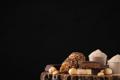 Different tasty energy bars, nuts and protein powder on wooden stump against black background, space for text