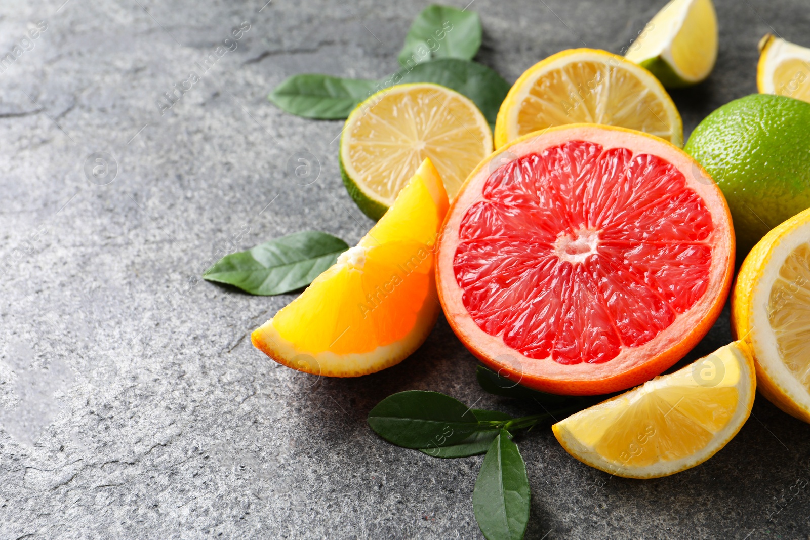 Photo of Different fresh citrus fruits and leaves on grey textured table, closeup. Space for text