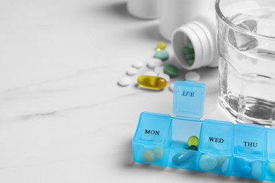 Weekly pill box with medicaments and glass of water on white marble table, closeup. Space for text