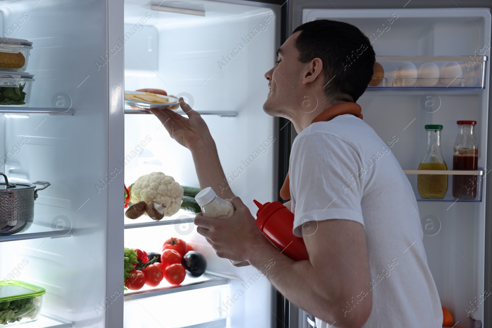 Photo of Happy man with sausages and sauces taking cheese out of refrigerator in kitchen at night