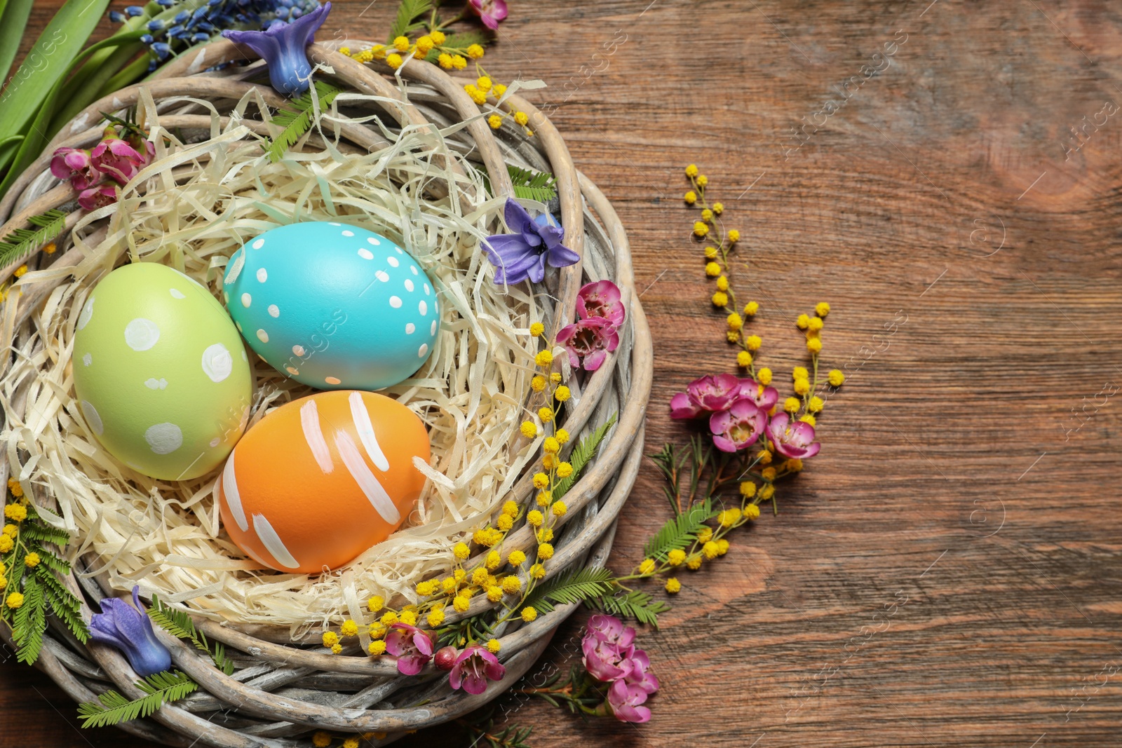 Photo of Flat lay composition of wicker nest with painted Easter eggs and flowers on wooden table, space for text