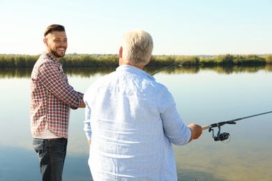 Father and adult son fishing together from riverside on sunny day