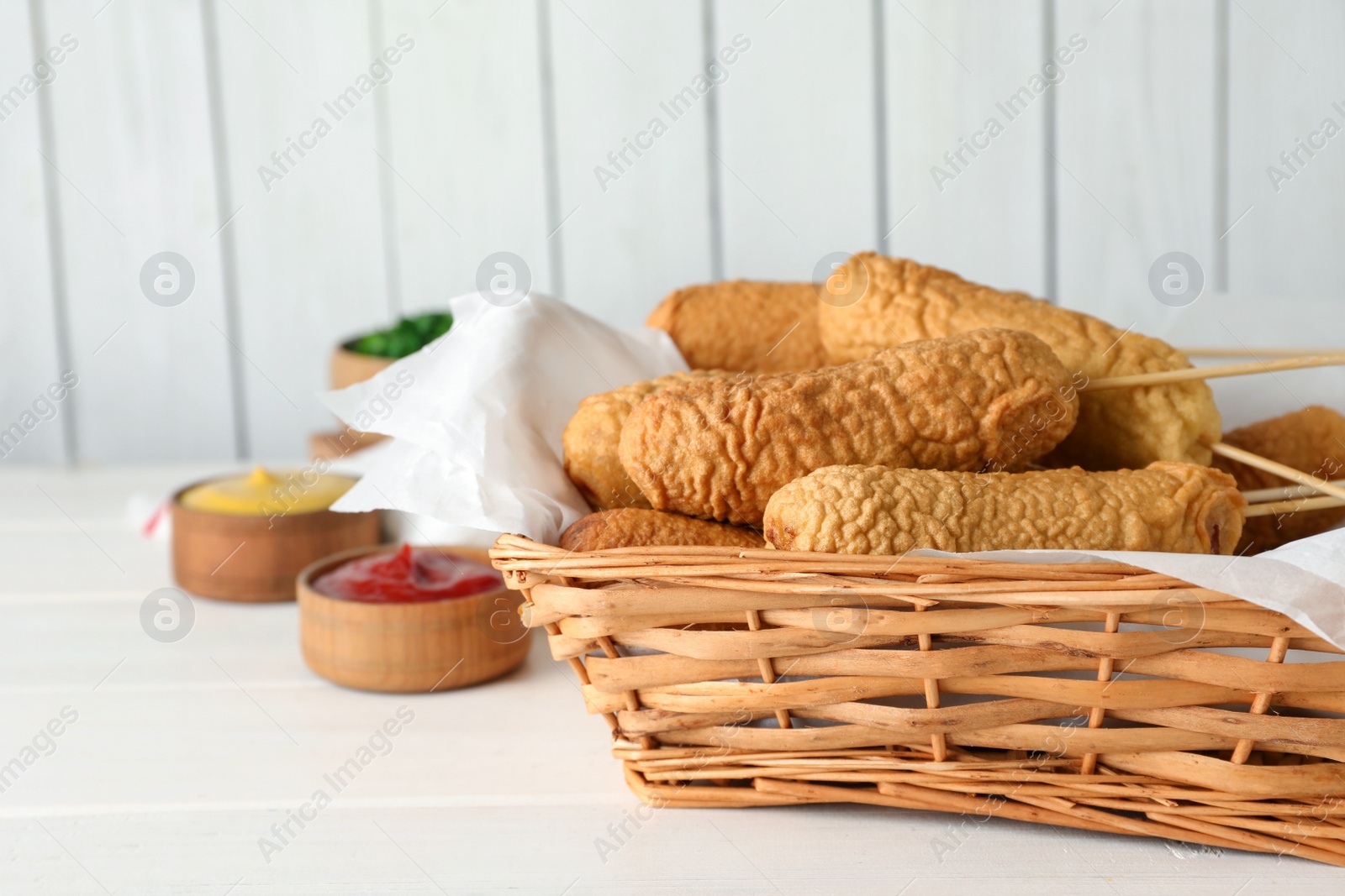 Photo of Delicious deep fried corn dogs in basket and sauces on white wooden table