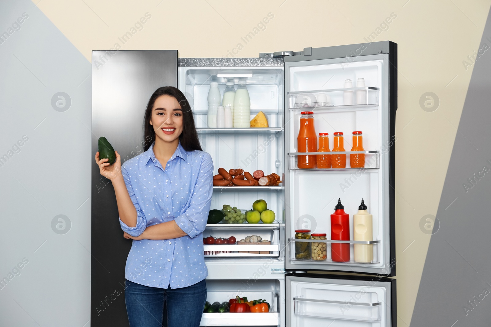 Photo of Happy young woman with avocado near open refrigerator indoors