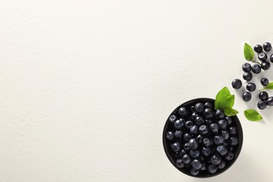 Tasty fresh bilberries with green leaves and bowl on white table, flat lay. Space for text