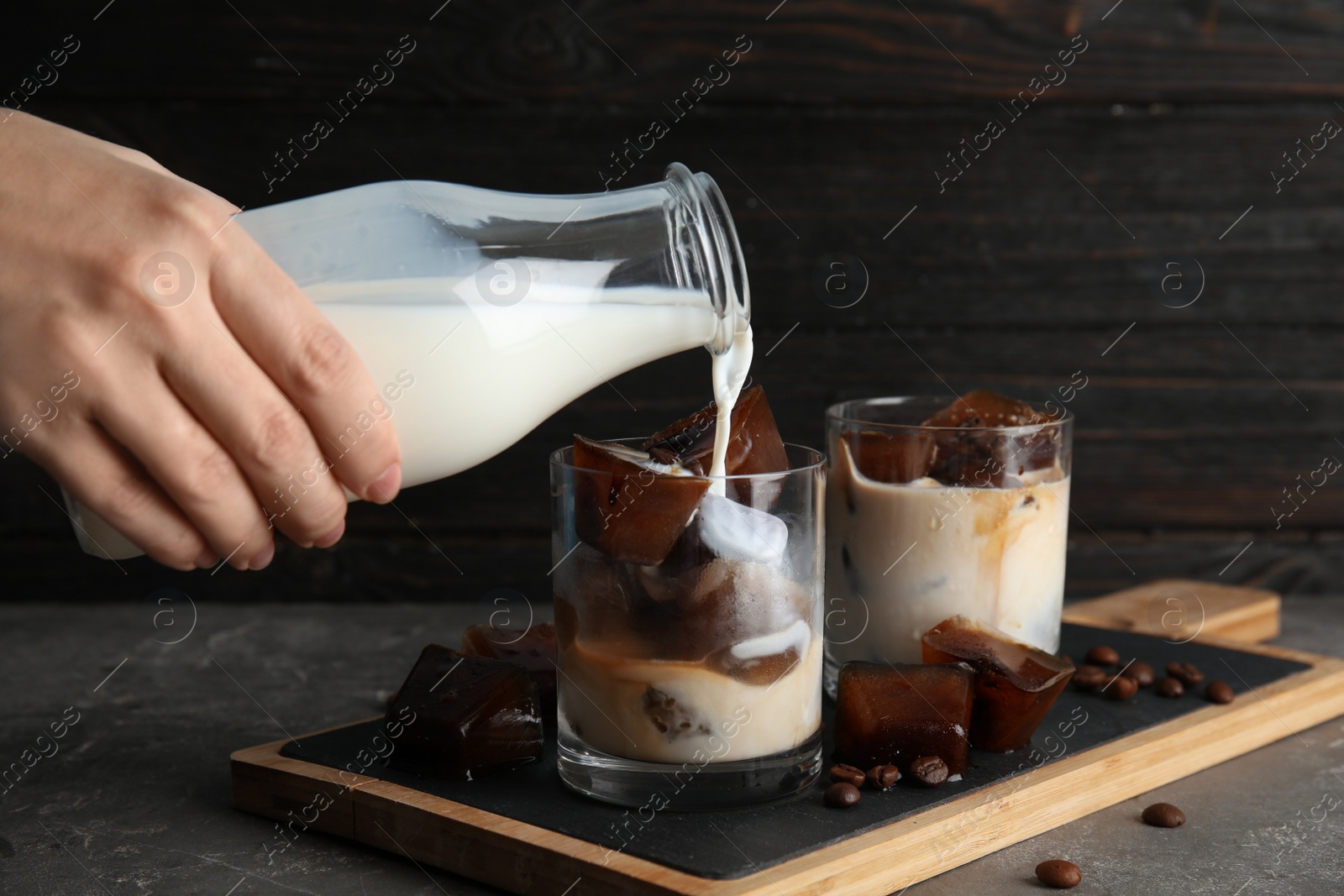 Photo of Woman pouring milk into glass with coffee ice cubes on table