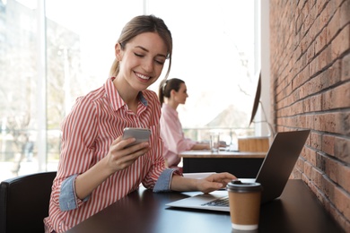 Young businesswoman with smartphone using laptop at table in office