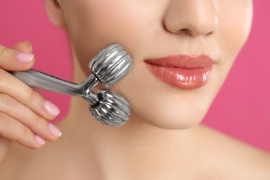 Woman using metal face roller on pink background, closeup