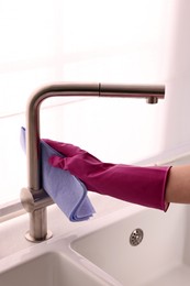 Photo of Woman in gloves cleaning faucet of kitchen sink with rag, closeup