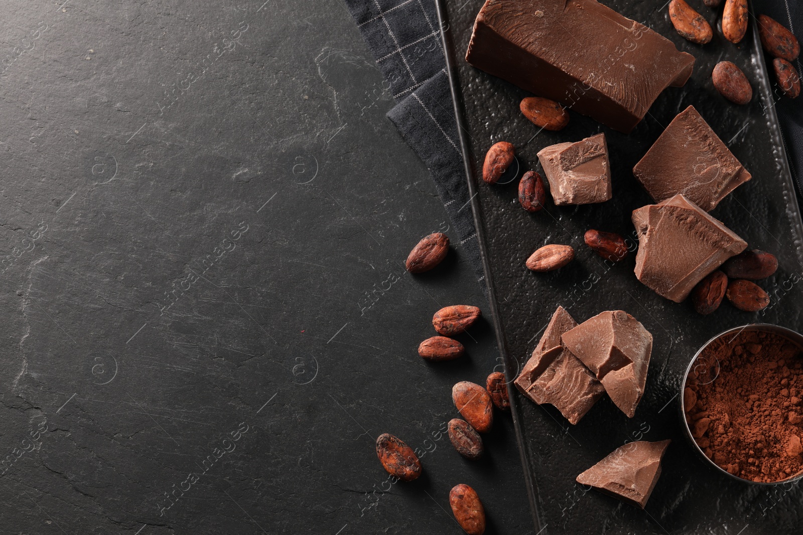 Photo of Pieces of tasty milk chocolate, cocoa beans and powder on black table, top view. Space for text