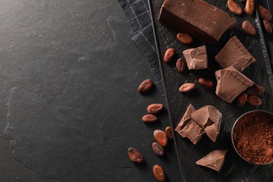 Pieces of tasty milk chocolate, cocoa beans and powder on black table, top view. Space for text