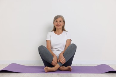 Photo of Happy senior woman sitting on mat near white wall. Yoga practice