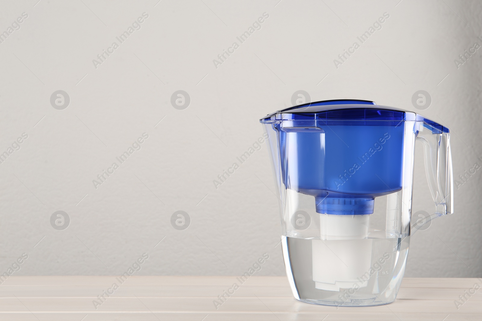 Photo of Filter jug with purified water on white table against light background. Space for text