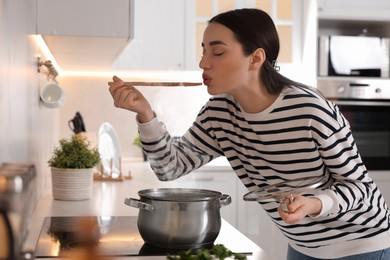 Photo of Beautiful woman with wooden spoon tasting soup in kitchen
