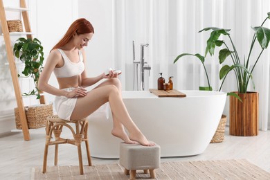 Beautiful young woman applying body cream onto legs in bathroom, space for text