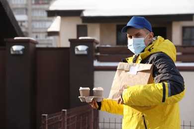 Photo of Courier in medical mask holding takeaway food and drinks near house outdoors. Delivery service during quarantine due to Covid-19 outbreak