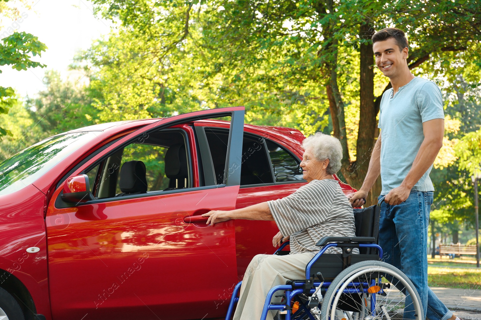 Photo of Young man helping disabled senior woman in wheelchair to get into car outdoors