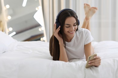 Photo of Young woman with smartphone and headphones listening to music on bed indoors. Space for text