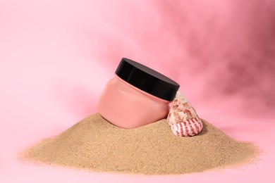 Jar of body cream and seashell on sand against pink background