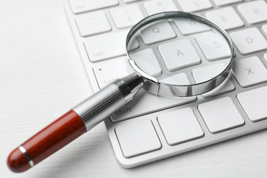 Magnifier glass and keyboard on white wooden table, closeup. Find keywords concept