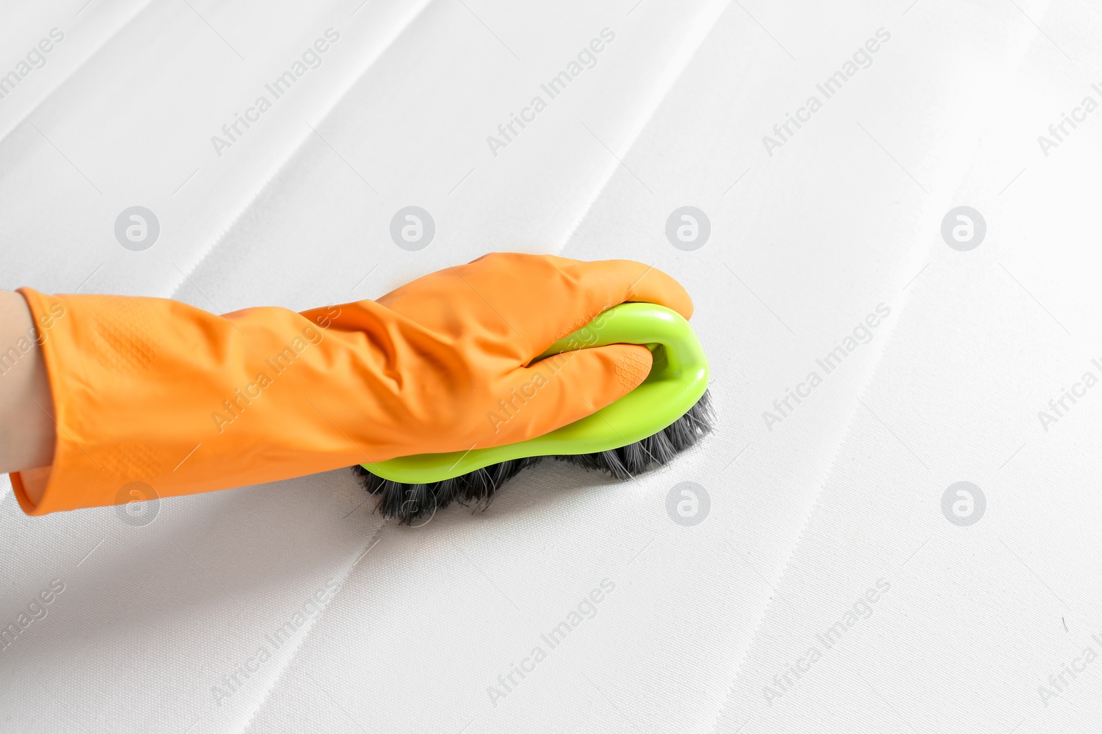 Photo of Woman in orange gloves cleaning white mattress with brush, closeup
