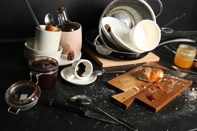 Many dirty utensils, dishware and food leftovers on black countertop. Mess in kitchen