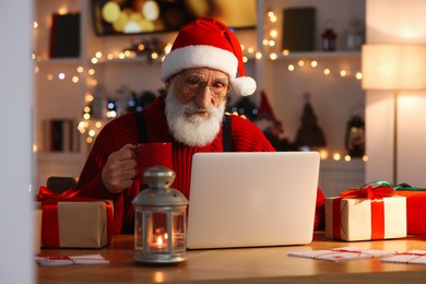 Photo of Santa Claus using laptop and drinking hot beverage at his workplace in room decorated for Christmas