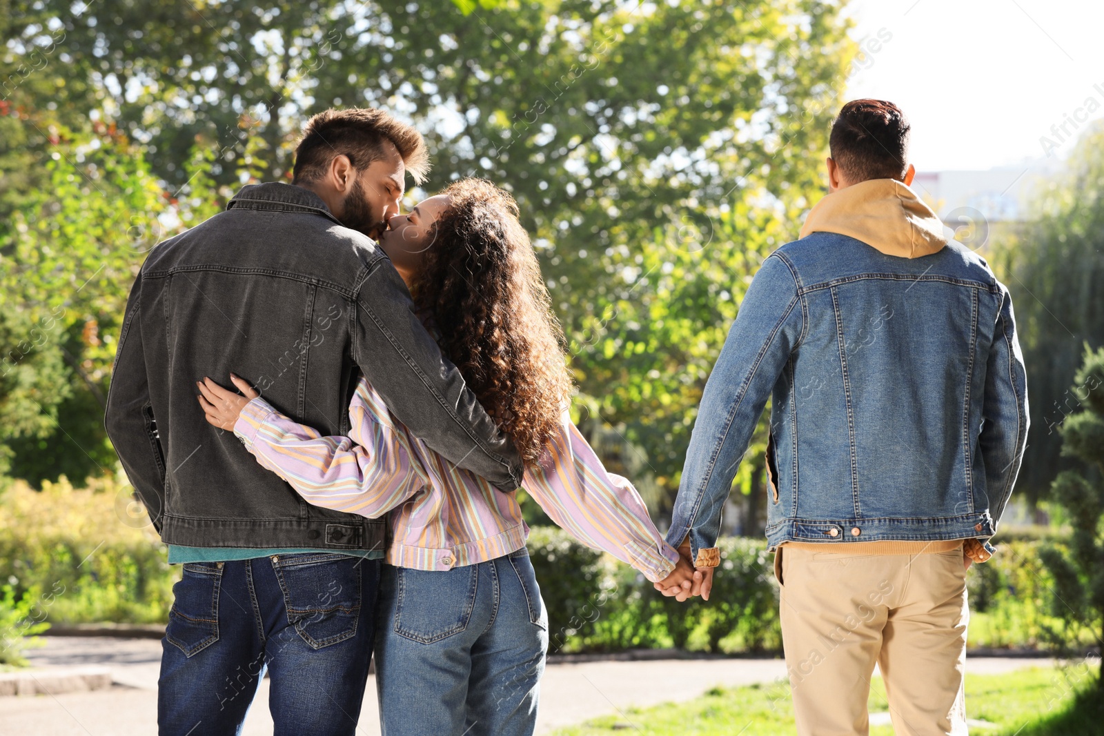 Photo of Woman kissing another man while holding hands with her boyfriend during walk in park, back view. Love triangle