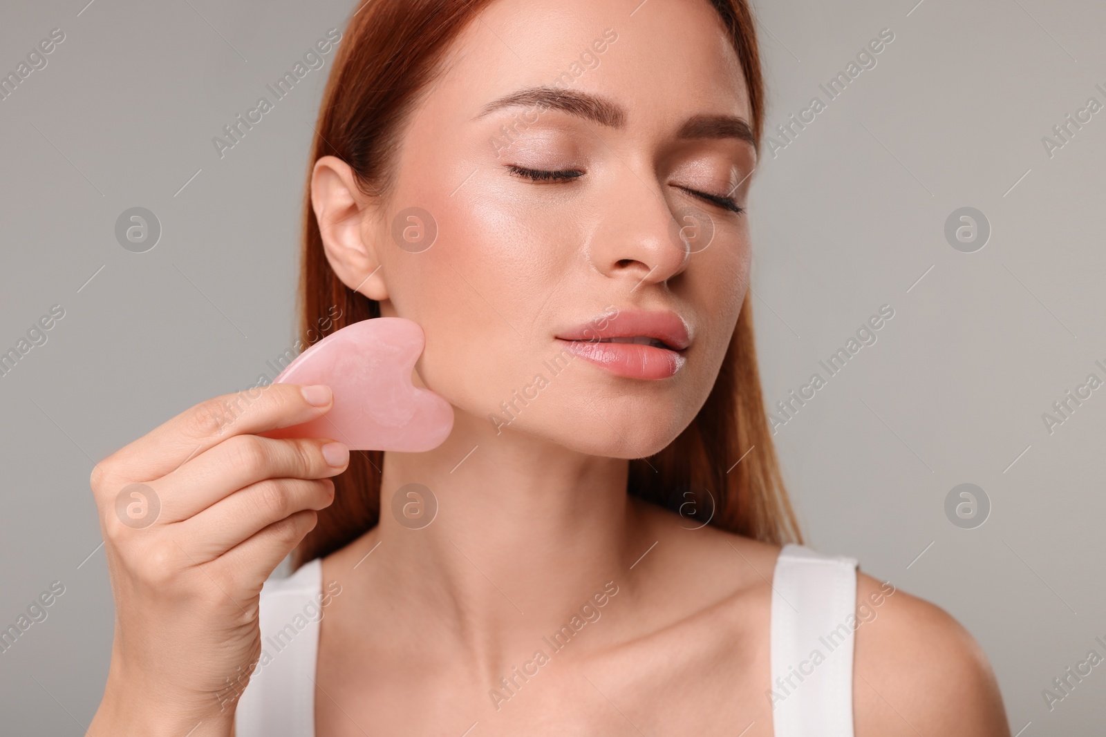 Photo of Young woman massaging her face with rose quartz gua sha tool on grey background