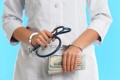 Photo of Doctor in handcuffs with bribe and stethoscope on light blue background, closeup. Corrupted medicine