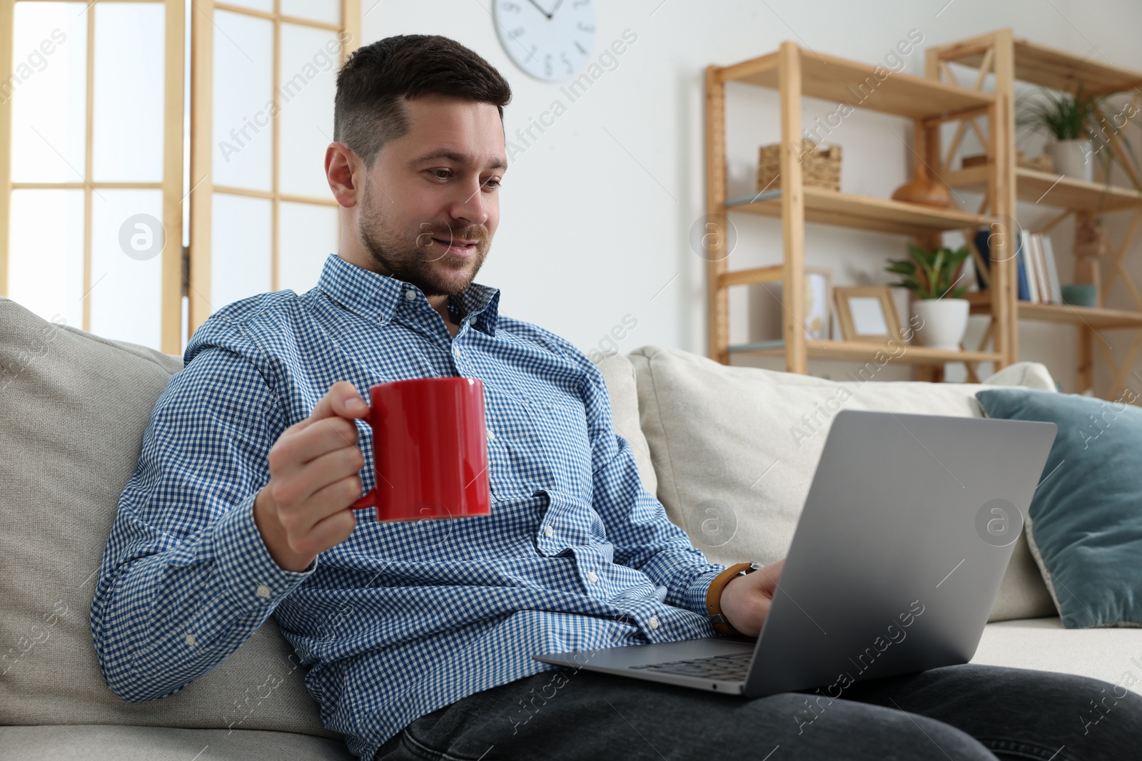 Photo of Happy man with cup of drink working with laptop on sofa at home