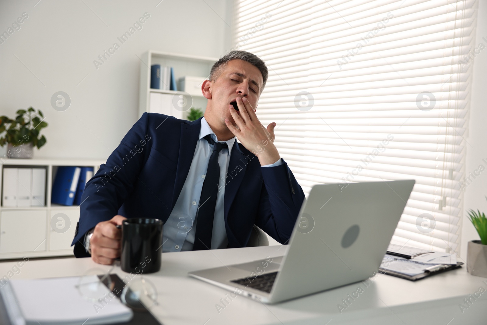Photo of Sleepy man with cup of drink yawning at table in office