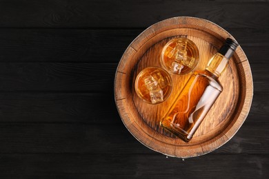 Photo of Whiskey with ice cubes in glasses, bottle and barrel on black wooden table, top view. Space for text