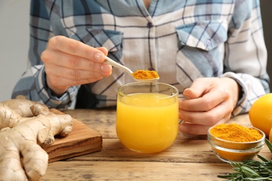 Photo of Woman adding turmeric to immunity boosting drink at wooden table with ingredients, closeup