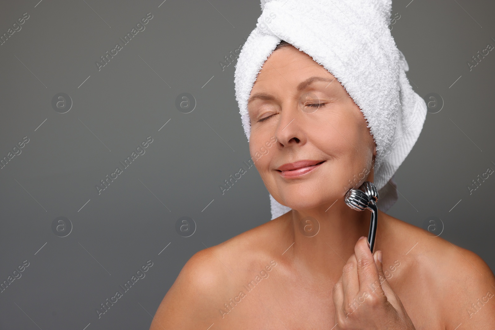 Photo of Woman massaging her face with metal roller on grey background, space for text
