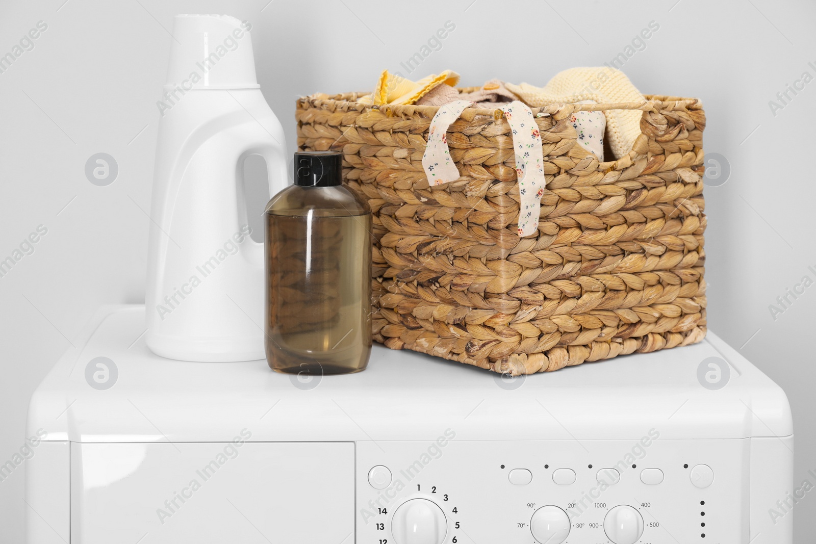 Photo of Baby clothes in wicker basket and laundry detergents on washing machine near light wall