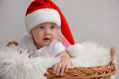 Photo of Cute baby wearing Santa hat in wicker basket on light grey background. Christmas celebration