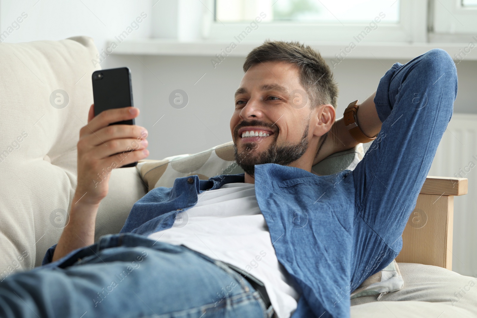 Photo of Happy man laying on sofa and using smartphone at home