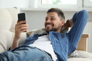 Photo of Happy man laying on sofa and using smartphone at home