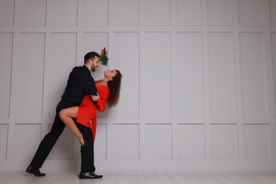 Lovely couple dancing under mistletoe bunch near light grey wall. Space for text