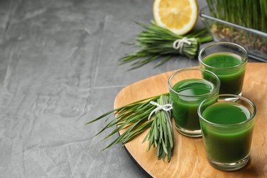 Photo of Wooden board with glasses of juice and fresh wheat grass on grey stone background. Space for text