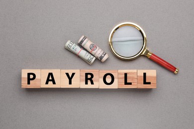 Word Payroll made of wooden cubes with letters, dollars and magnifying glass on grey background, flat lay