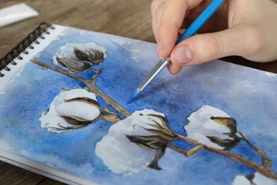 Photo of Woman painting cotton flowers in sketchbook at table, closeup