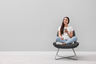 Photo of Beautiful woman with smartphone sitting in armchair near light grey wall indoors, space for text