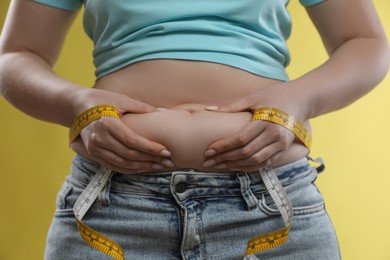 Photo of Woman with measuring tape touching belly fat on yellow background, closeup. Overweight problem