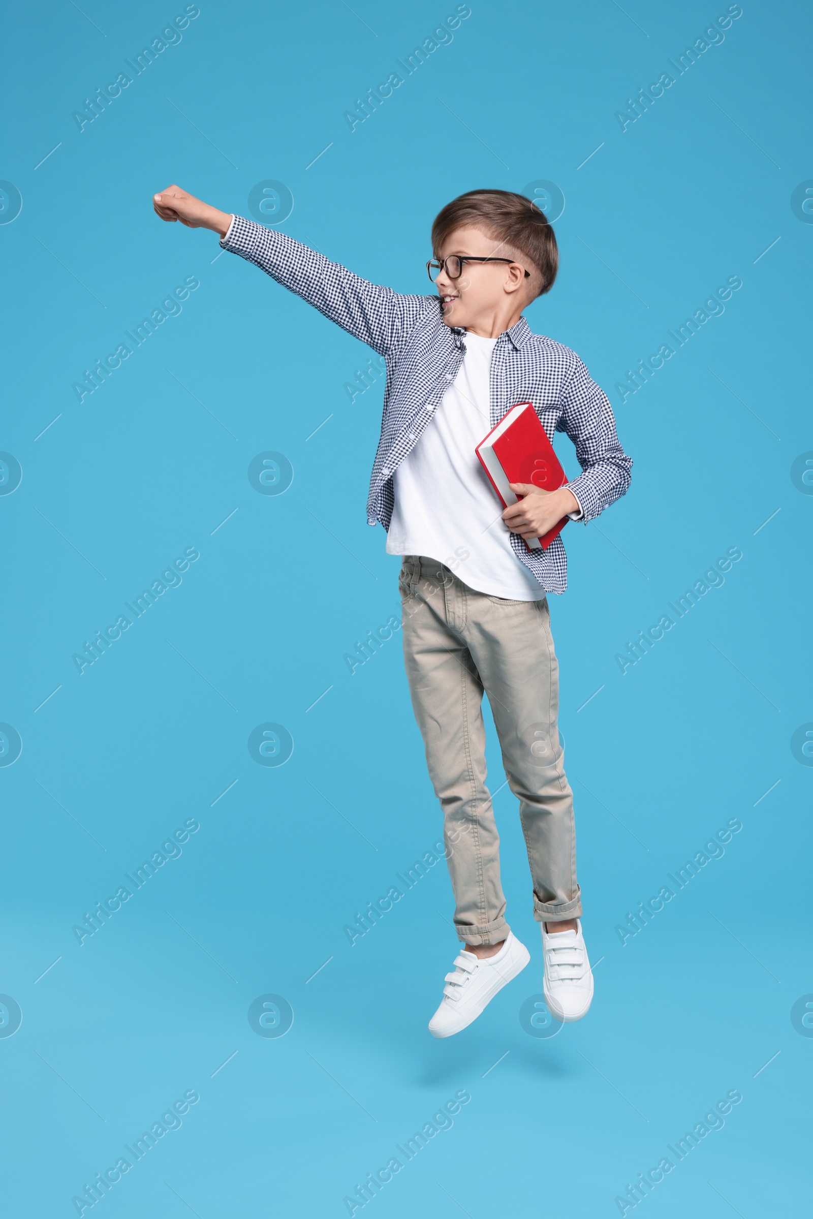 Photo of Cute schoolboy in glasses holding book and jumping on light blue background