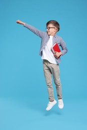 Cute schoolboy in glasses holding book and jumping on light blue background