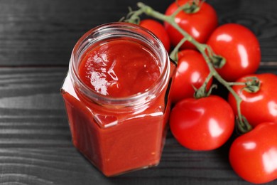 Photo of Delicious ketchup in jar and tomatoes on black wooden table, closeup. Tomato sauce