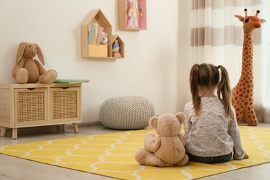 Photo of Lonely little girl with teddy bear sitting on floor at home, back view. Autism concept