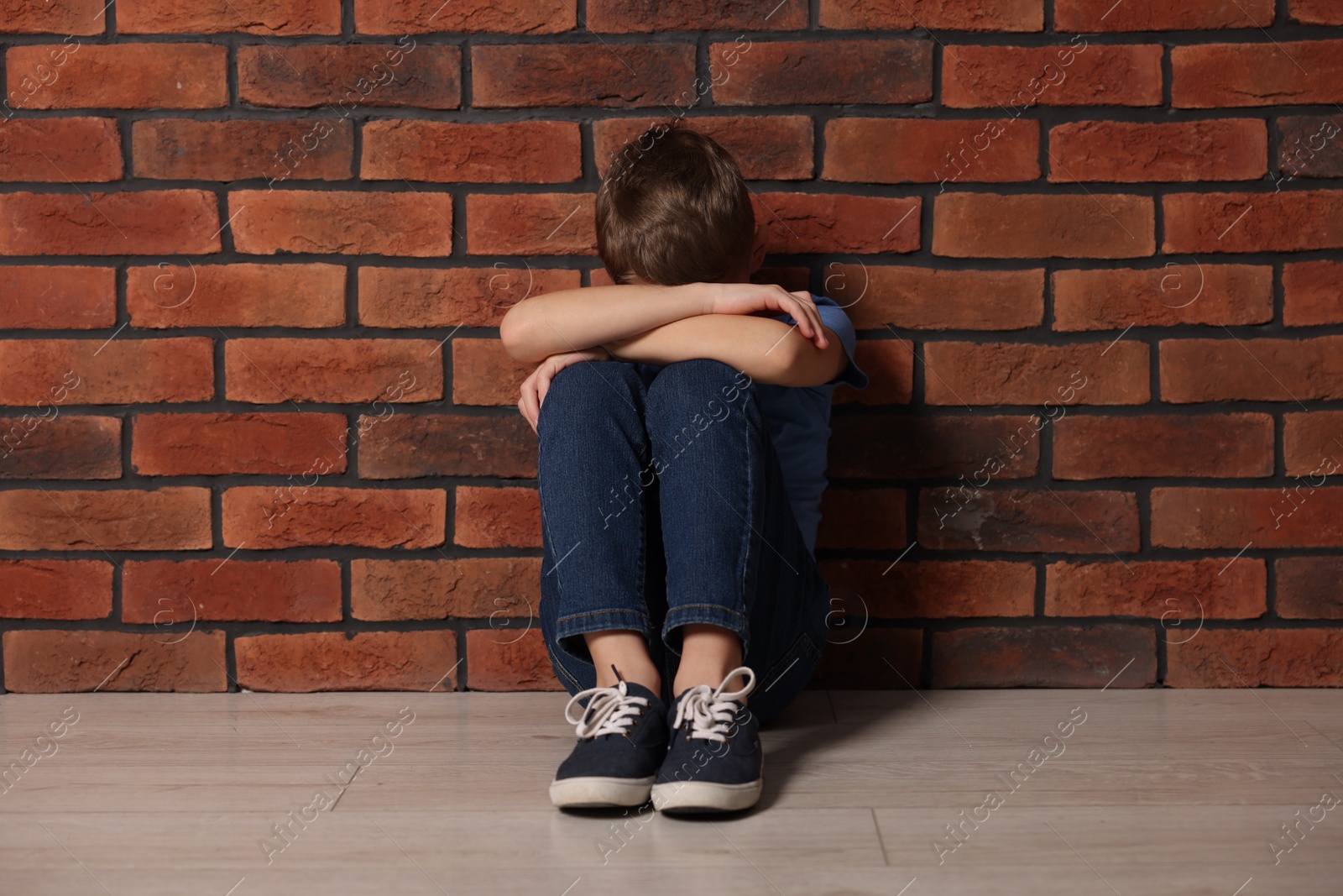 Photo of Child abuse. Upset boy sitting on floor near brick wall indoors